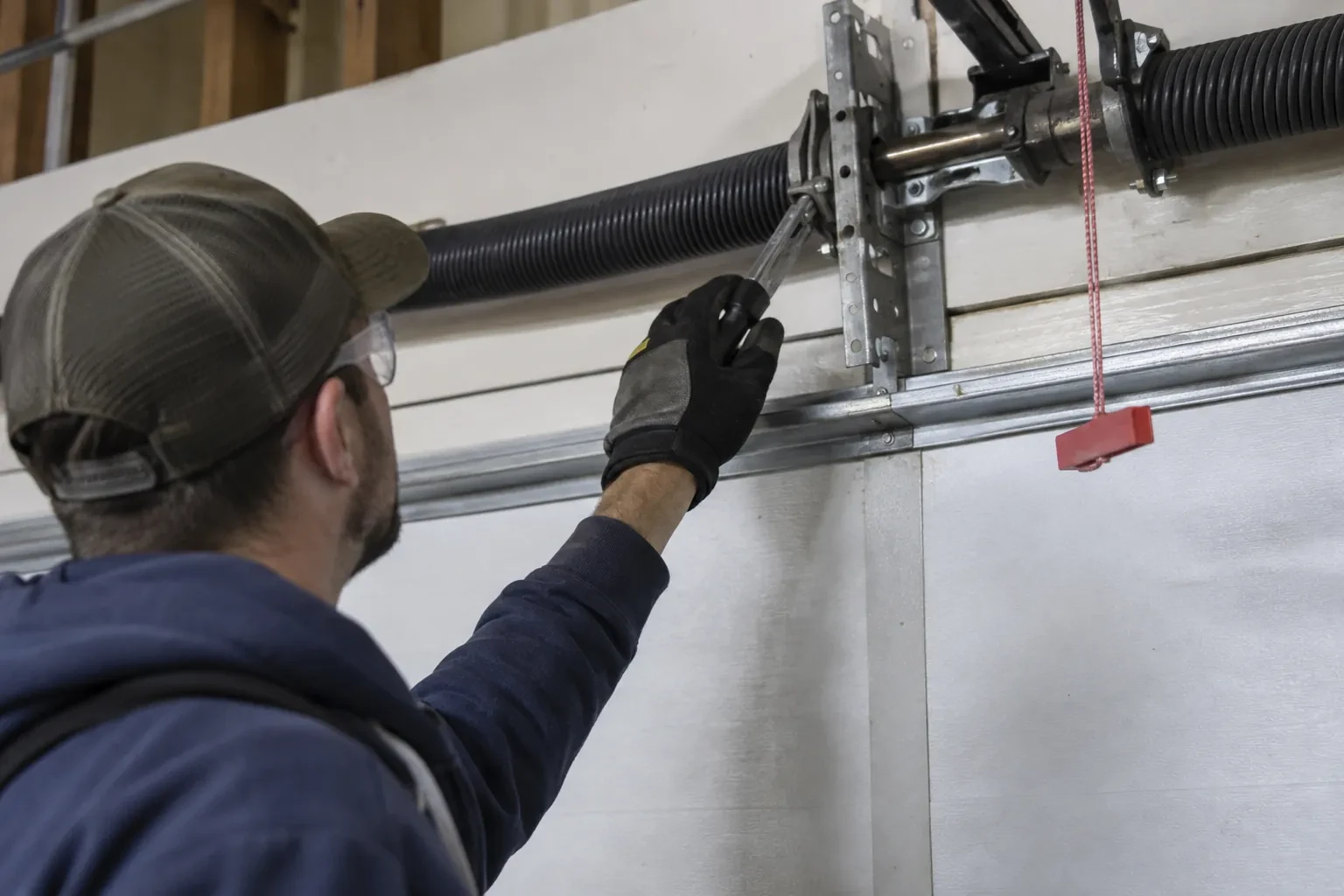 Technician in a cap reaching a gloved hand with a tool toward a coiled spring and bracket above a garage door. — Toronto