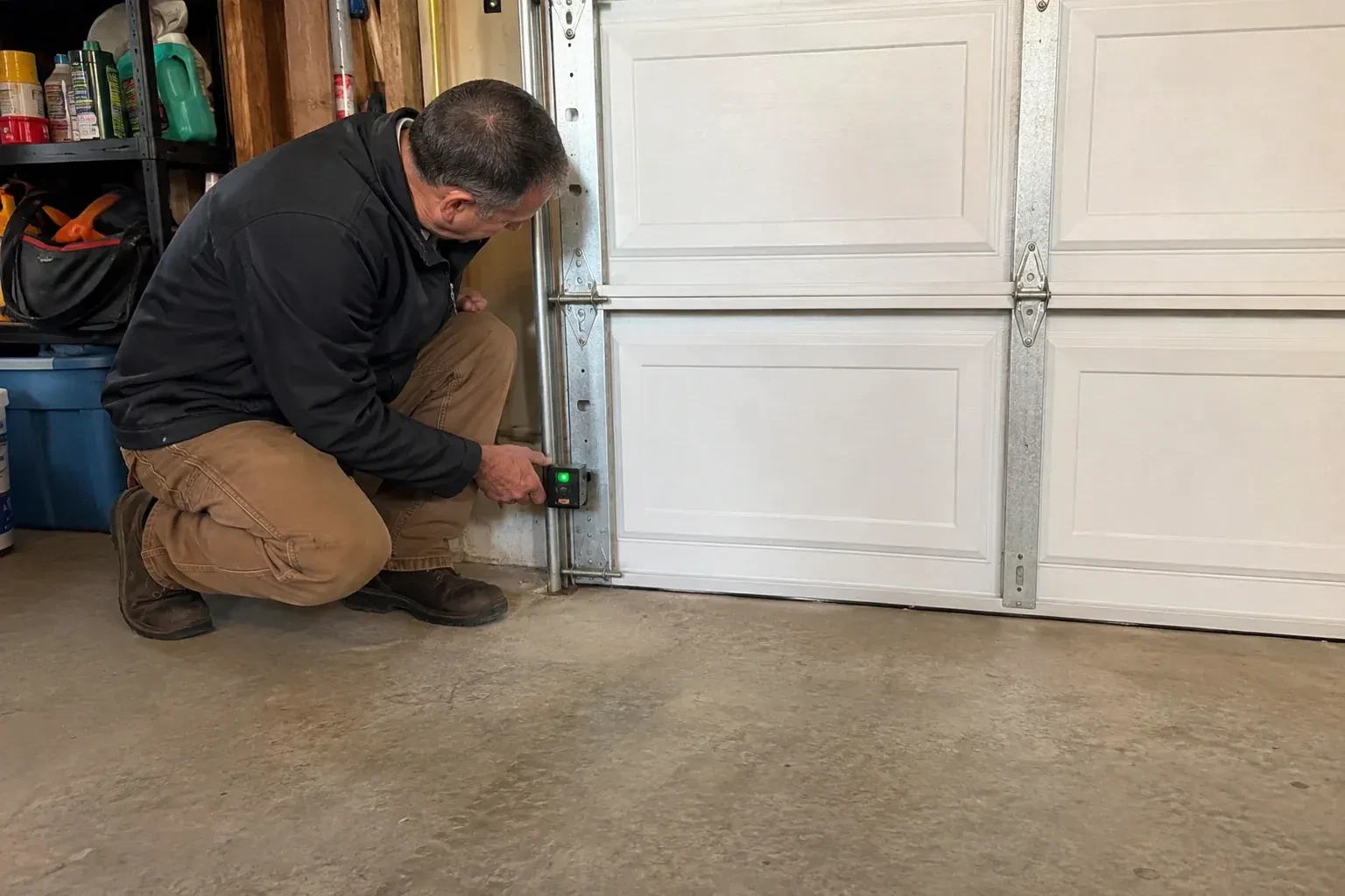 Technician kneeling beside a white paneled garage door, holding a small black box with a green light against the vertical track. — Photo-Eye Sensor Alignment or Replacement, Toronto